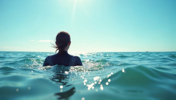 Back view of a woman wearing a wetsuit swimming in the ocean, with waves splashing under a bright, clear sky. photo