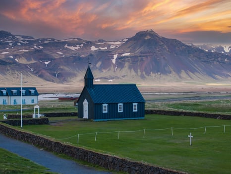 A black church stands in a green field with a mountain range in the background. The sky is a beautiful pink and orange color, and a small river is visible in the distance. photo