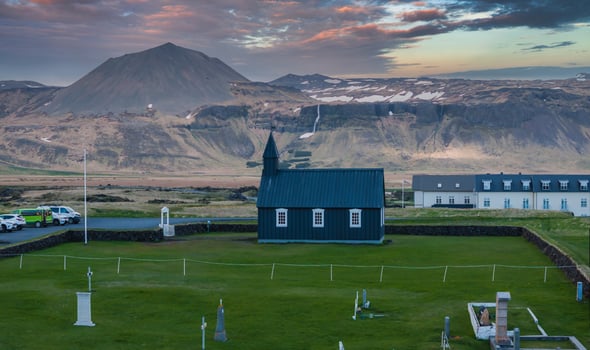 A small black church with white trim stands in a grassy field, surrounded by a stone wall, with snow-covered mountains and a waterfall in the background. photo