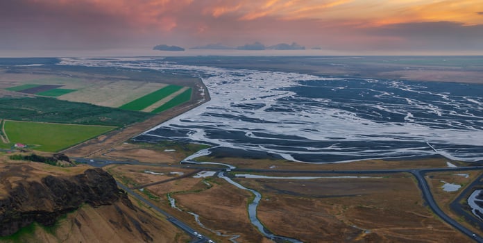 Stunning aerial view of a vast, winding river in Iceland, surrounded by a mix of brown, green, and black terrain. Small buildings are visible in the distance. photo