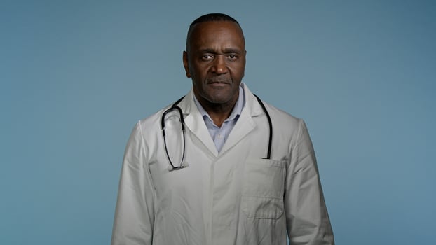 Mature confident happy doctor standing in gray background studio with arms crossed glad smiling african american man professional therapist physician in medical gown posing indoors looking at camera photo