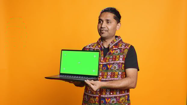Tech content creator doing influencer marketing using green screen laptop, isolated over studio background. Indian man holding empty copy space mockup notebook, doing review, camera B photo