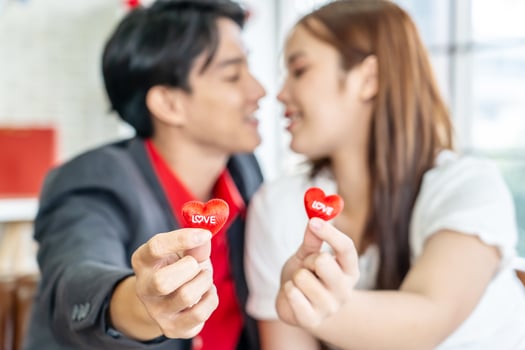Happy Asian couple holding red hearts while sitting on sofa in living room with red heart balloons, romantic lover celebrating anniversary on Valentine Day together, selective focus photo