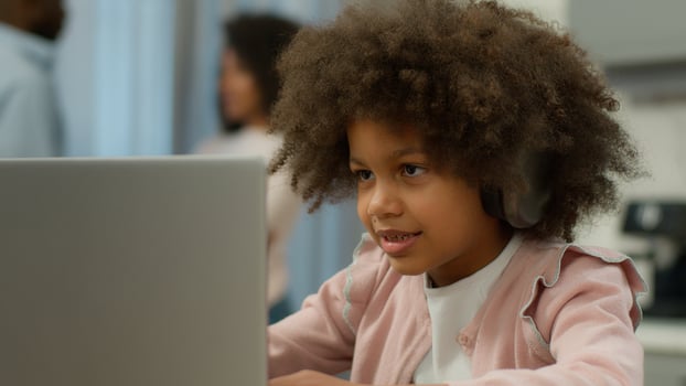 Blurred mother father parents talking background concentrated African American little girl daughter child kid in headphones laptop e-learning online class study typing play game family in kitchen home photo