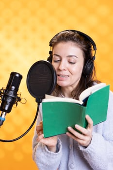 Smiling narrator wearing headphones reading aloud from book into microphone against yellow background. Cheerful person recording audiobook, creating engaging media content for listeners photo