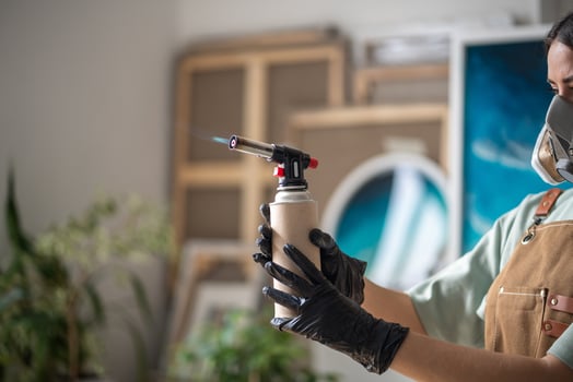 Woman with respirator ignites blowtorch, adjusts flame in artist workshop opposite epoxy paintings photo