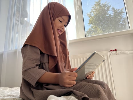 Asian Muslim little girl wearing hijab sitting near window while streaming movies on a tablet photo