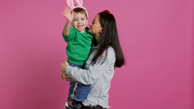Mother lifting up her little boy and kissing him in studio, showing love and hugging cute kid against pink background. Sweet mom and son embracing each other and laughing. Camera B. photo