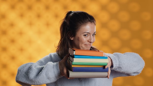 Cheerful woman holding stack of books arriving in studio with yellow backdrop, smiling. Joyous person walking with pile of novels, feeling delighted and eager to read, camera A photo