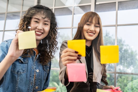 Asian creative business women are writing note on the text sheet after thinking new idea and planning project with colleague, Two employee brainstorming idea on glass wall and sticky photo