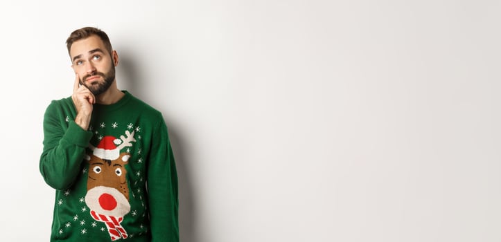 New Year, holidays and celebration. Sad young man with beard, wearing green sweater, showing a teardrop on his face and looking up upset, standing over white background photo