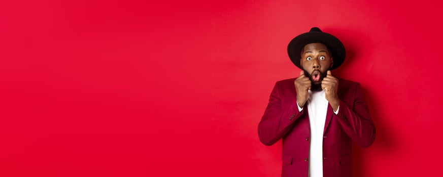 Fashion and party concept. Surprised african american male model looking shocked and amazed, saying wow and staring camera, standing against red background photo