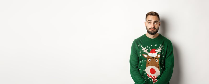 New Year, holidays and celebration. Gloomy bearded man in sweater, looking indecisive and looking aside, standing against white background photo
