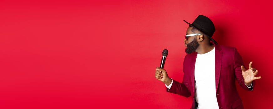 Passionate black male singer performing against red background, singing into microphone, wearing party outfit, standing over red background photo