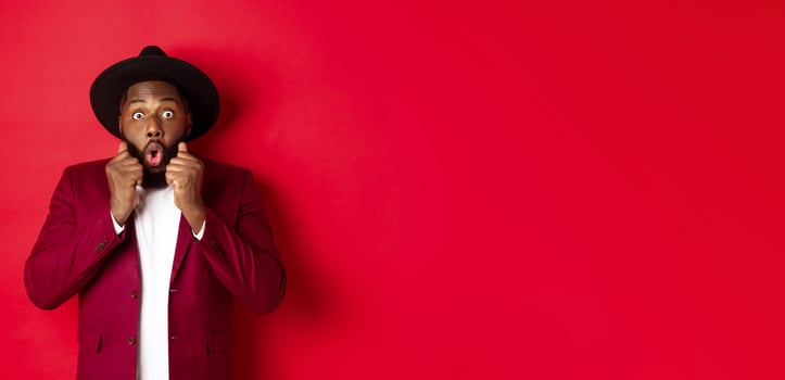 Fashion and party concept. Surprised african american male model looking shocked and amazed, saying wow and staring camera, standing against red background photo