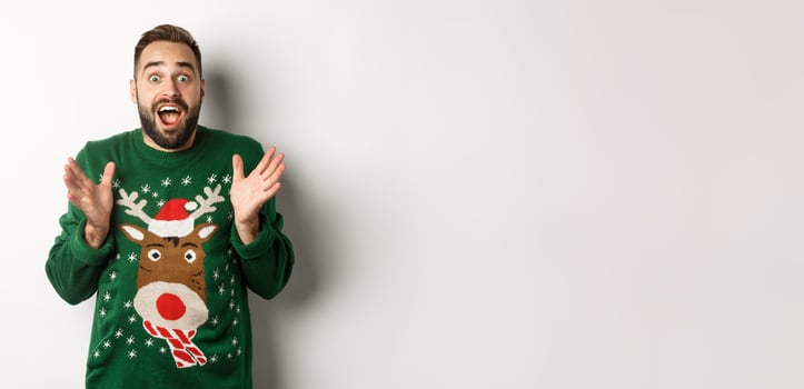 New year celebration and winter holidays concept. Happy and surprised bearded guy looking amazed, catching something, standing in funny Christmas sweater, white background photo