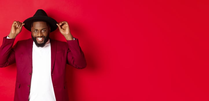 Fashion and party concept. Handsome african american man wearing hat and jacket, smiling self-assured, standing against red background photo