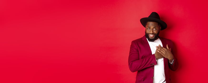 Fashion and party concept. Handsome african american macho man flirting, smiling and holding hands on heart, being grateful and touched, standing over red background photo