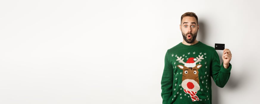 Christmas holidays and shopping concept. Surprised handsome man in sweater showing credit card, standing over white background photo