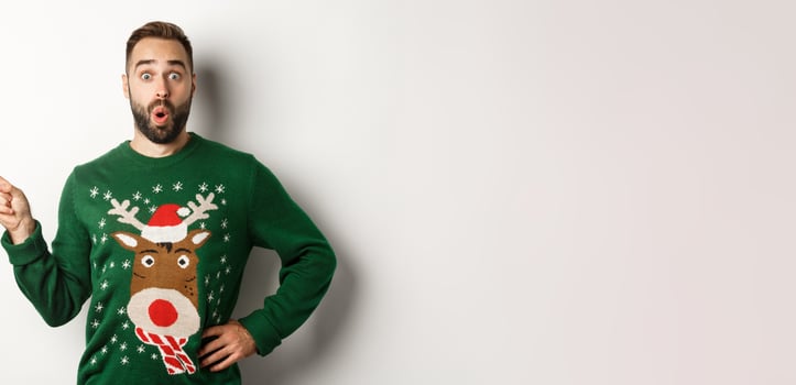 New year celebration and winter holidays concept. Surprised man in christmas sweater pointing finger at upper left corner, looking amazed, white background photo