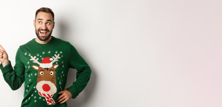 New year celebration and winter holidays concept. Dreamy bearded man in green christmas sweater, pointing at upper left corner and smiling amused, white background photo