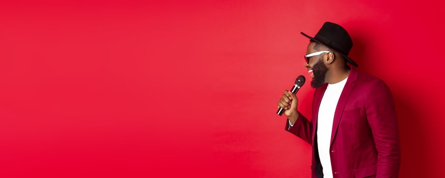 Passionate black male singer performing against red background, singing into microphone, wearing party outfit, standing over red background photo