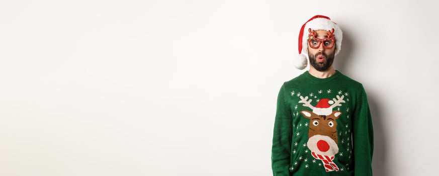 Christmas holidays, celebration concept. Surprised guy in party glasses and santa hat looking down amused, standing over white background photo