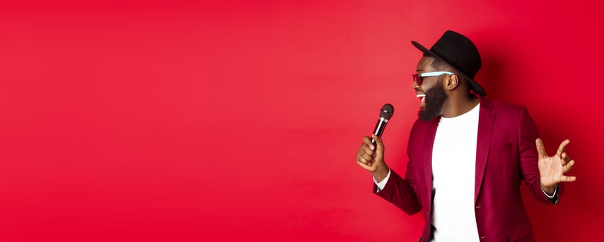 Passionate black male singer performing against red background, singing into microphone, wearing party outfit, standing over red background photo