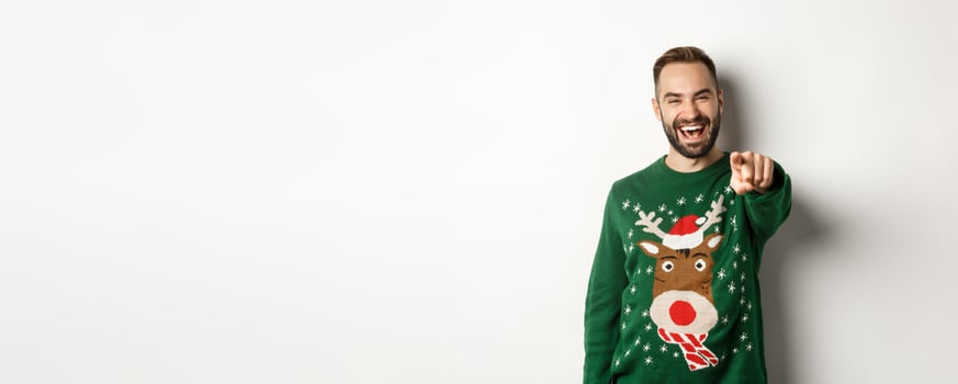 Winter holidays and christmas. Happy young man celebrating New Year eve, pointing finger at you and laughing, standing over white background photo