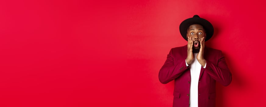 Fashion and party concept. Impressed Black man in classy outfit staring with complete disbelief at camera, gasping and looking surprised, standing over red background photo