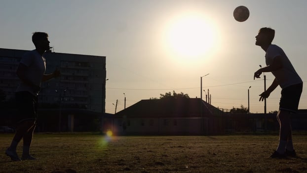 Silhouette of professional footballers kicking soccer ball with their heads on stadium at sunset. Two sportsmen showing tricks with ball while passing it to each other at field. Freestyle football photo