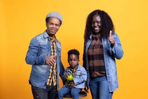 African american mom and dad giving like symbol in front of camera, posing with their adorable son sitting on the chair. Joyful young parents showing thumbs up sign, approving in studio. photo