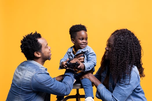 Proud young parents playing with their adorable son in studio, hugging him against yellow background. African american mom and dad posing with their small child holding a controller. photo