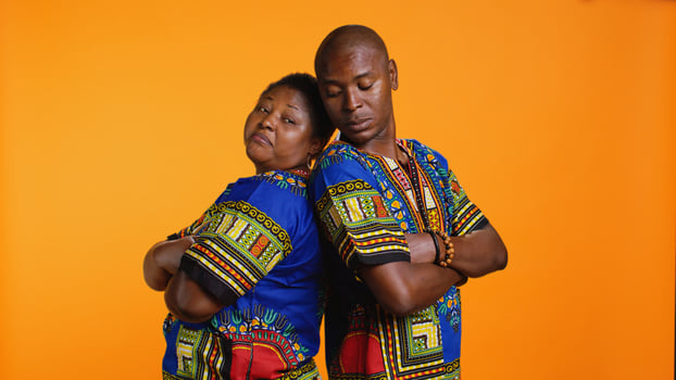 Serious proud ethnic couple standing back to back in studio, posing with confidence over orange background. African american man and woman dressed in traditional clothing, carefree partners. photo