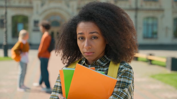 Sad upset stressed disappointed African American student girl female woman lady teen teenager standing alone in city outdoors university college high school suffer bullying talking students background photo