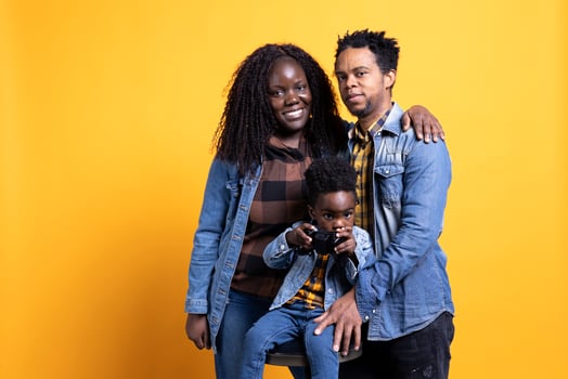 Joyful little child playing with a controller for games, posing with his mother and father in studio. African american family with a toddler standing against yellow background, kid gaming. photo