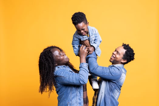 Loving african american parents lifting their cute small son in studio, posing in front of the camera. Proud happy couple bonding with their adorable toddler against yellow background. photo
