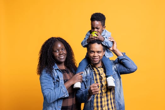 Portrait of african american parents posing with their child on camera, feeling happy and grateful with their little toddler. Adorable small boy eating an apple on his fathers shoulders. photo