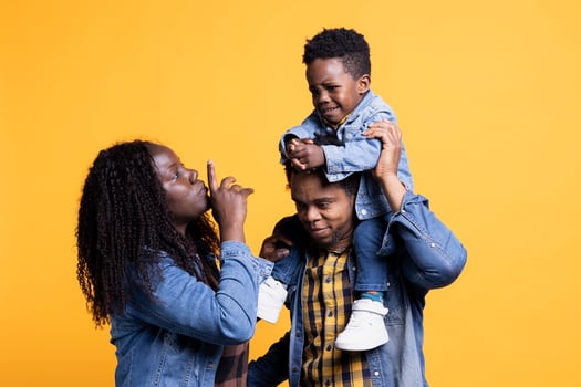 African american parents calming down their infant son while he cries, his father is carrying him on his shoulders. Young mother showing hush sign to his toddler, trying to make him smile. photo