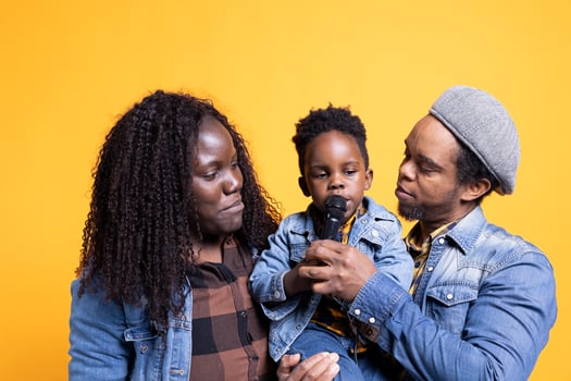 African american couple making their toddler sing in front of the camera, using microphone and having fun together. Loving parents listening to their child singing in studio, talented young boy. photo