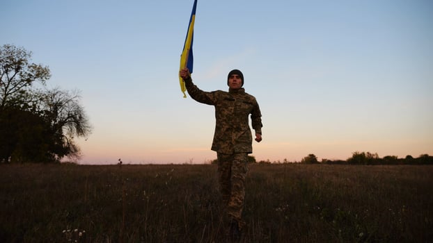 Soldier of ukrainian army running with raised blue-yellow banner on field at dusk. Young male military in uniform jogging with flag of Ukraine at meadow. Victory against russian aggression concept photo