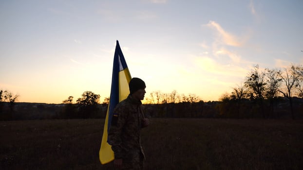Young male military in uniform walks with flag of Ukraine at meadow. Soldier of ukrainian army going with blue-yellow banner on field at sunset. Concept of victory against russian aggression. Slow mo photo