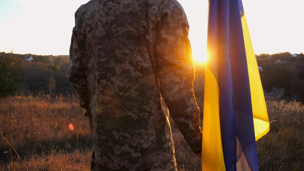 Male ukrainian army soldier stands with national banner against background of beautiful sunset. Young man in camouflage uniform with Ukraine flag as symbol of victory against russian aggression photo
