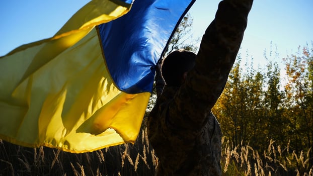 Military man in uniform stands with raised over head flag of Ukraine at countryside. Male ukrainian army soldier with lifted national banner in honor of victory against russian aggression. End of war. photo