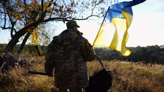 Young male military in uniform walks with waving flag of Ukraine at countryside. Soldier of ukrainian army going with blue-yellow banner outdoor. Concept of victory against russian aggression. photo