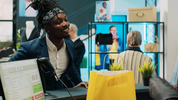 Retail clerk putting clothes in a paper bag for customer, working at cash register and selling formal fashion items at department store. Woman employee assisting african american client. Camera A. photo