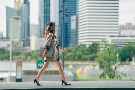A young professional woman walking confidently in an urban setting with a city skyline and park in the background, holding a tablet, dressed in business casual attire, showing determination photo
