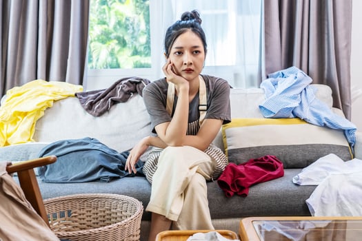 Stressed young woman asian sitting on the sofa in the living room and among a mess at home, woman sitting among scattered clothes on sofa and ignoring, woman frowning and letting out soft sigh. photo