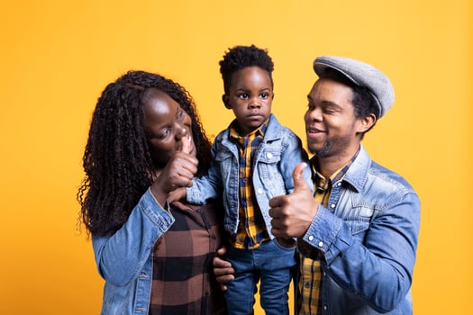 Cheerful african american mom and dad showing thumbs up sign and holding their infant son, grateful family bonding against yellow background. Positive husband and wife give like symbol. photo