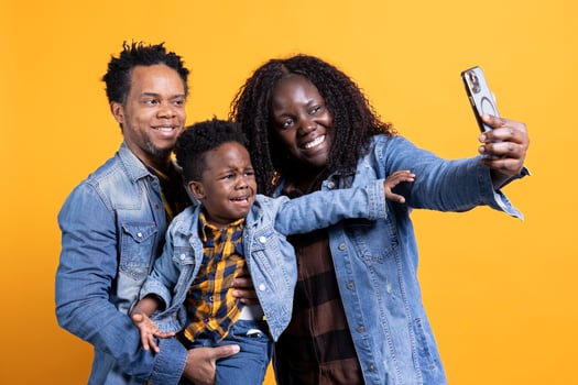 African american mom and dad taking pictures with their small kid against yellow background, making sweet funny memories together. Happy parents posing for photos with the toddler.